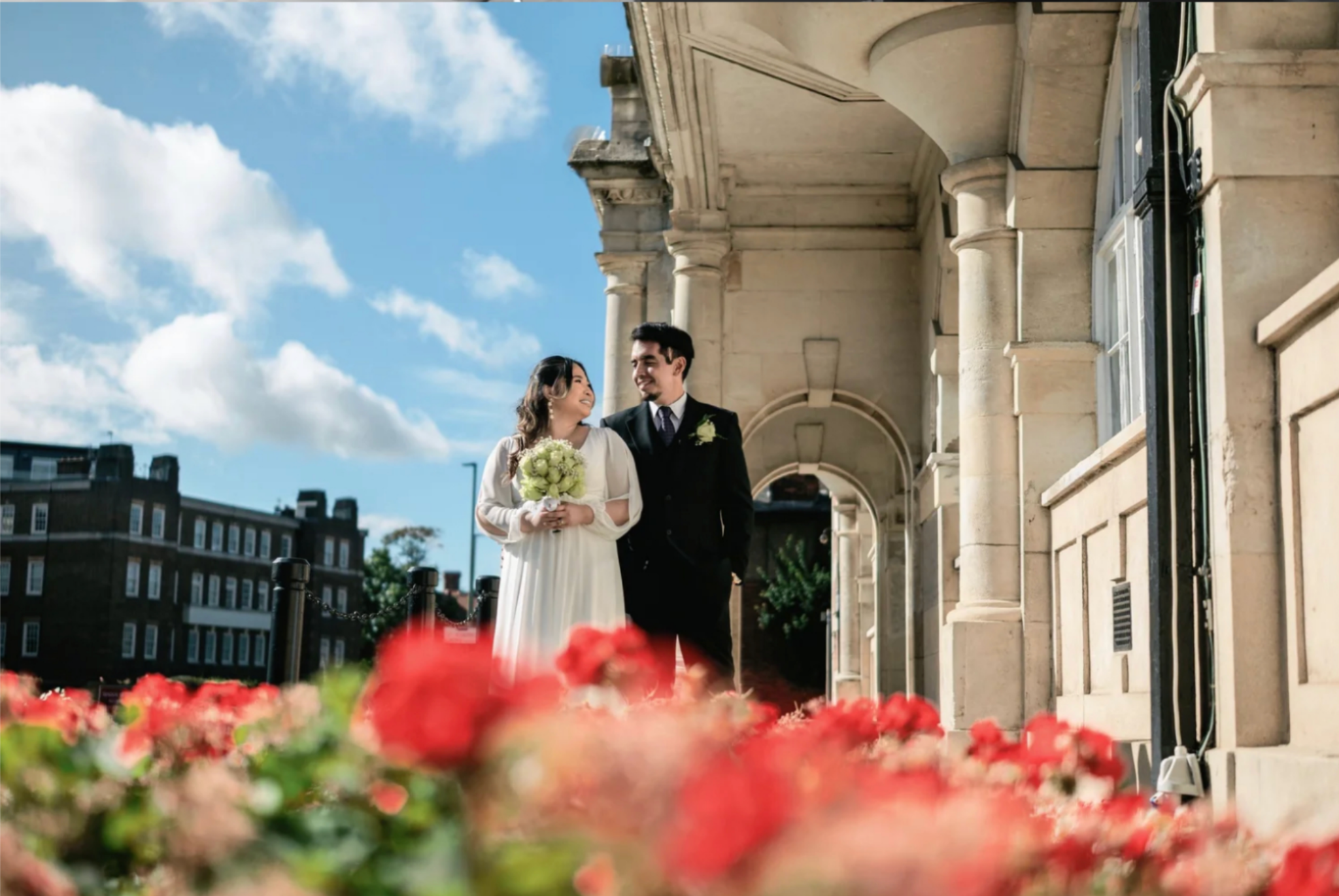 Newlyweds walking beside a city building and red flowers
